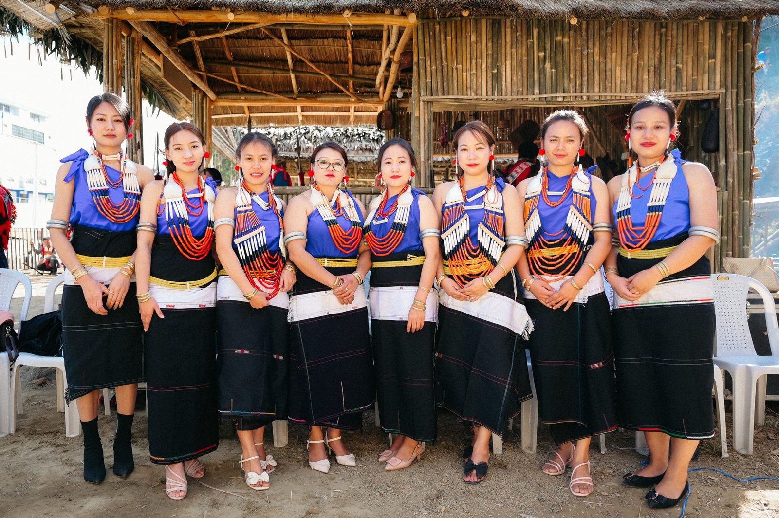 Eight women in Rengma traditional dress standing before a bamboo and thatch structure