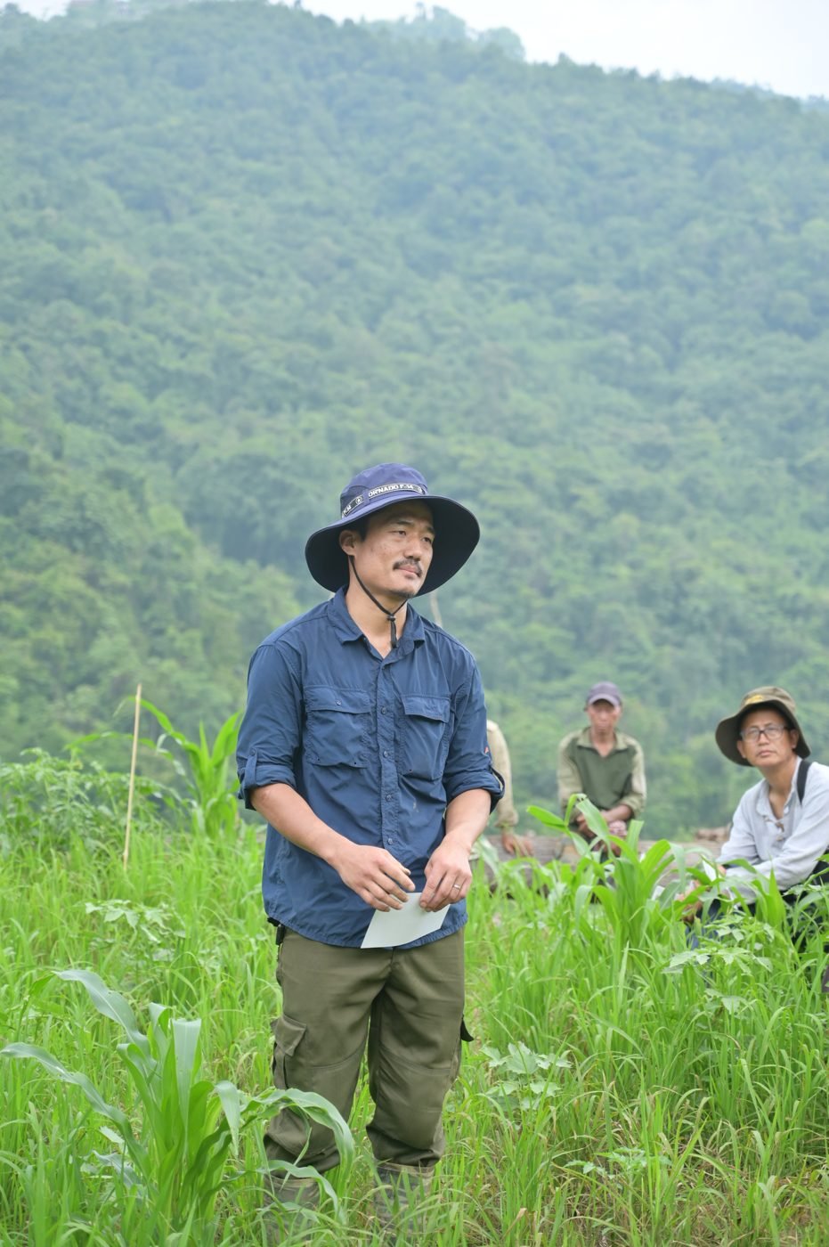 Ruchinilo Kemp in a wide-brimmed hat standing in tall grass, forested hills behind him