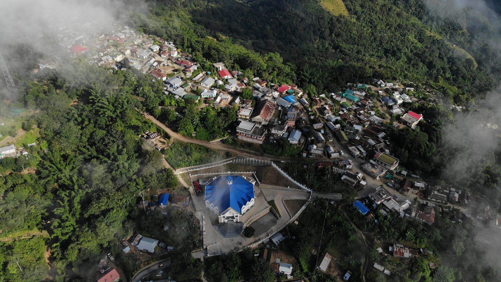 A village on a forested slope with a blue-roofed church, seen through low cloud